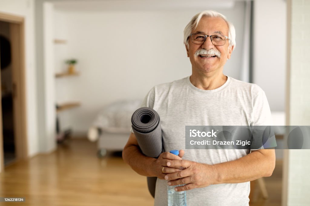 Happy senior man holding bottle of water and exercise mat in the living room and looking at camera.